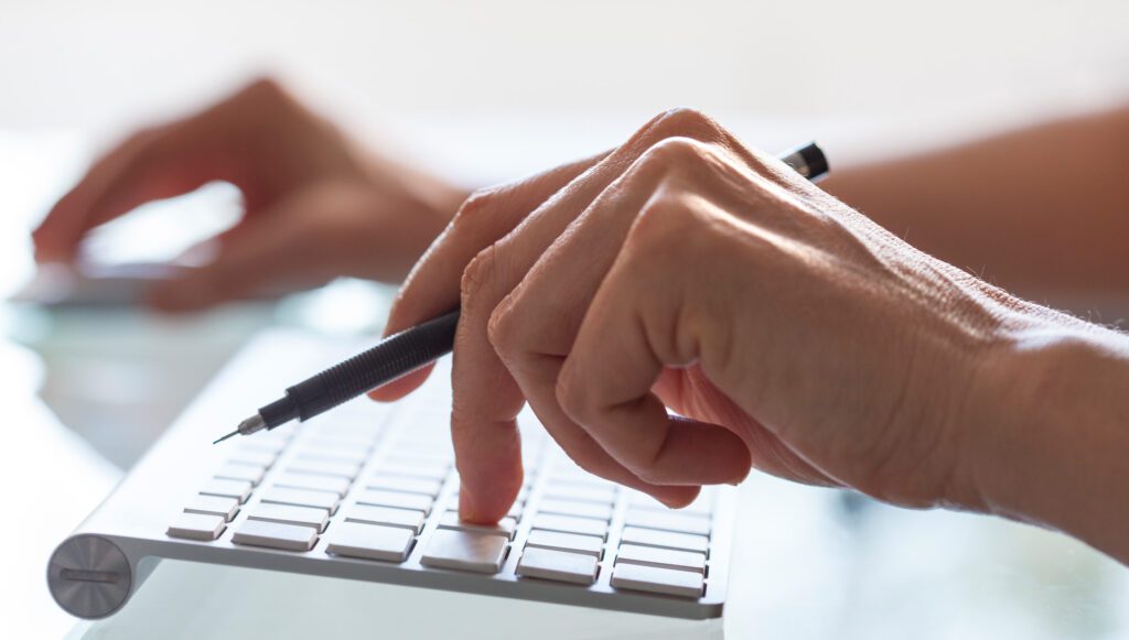 Close-up of hands typing on a keyboard while holding a pen, with another hand using a mouse in the background.