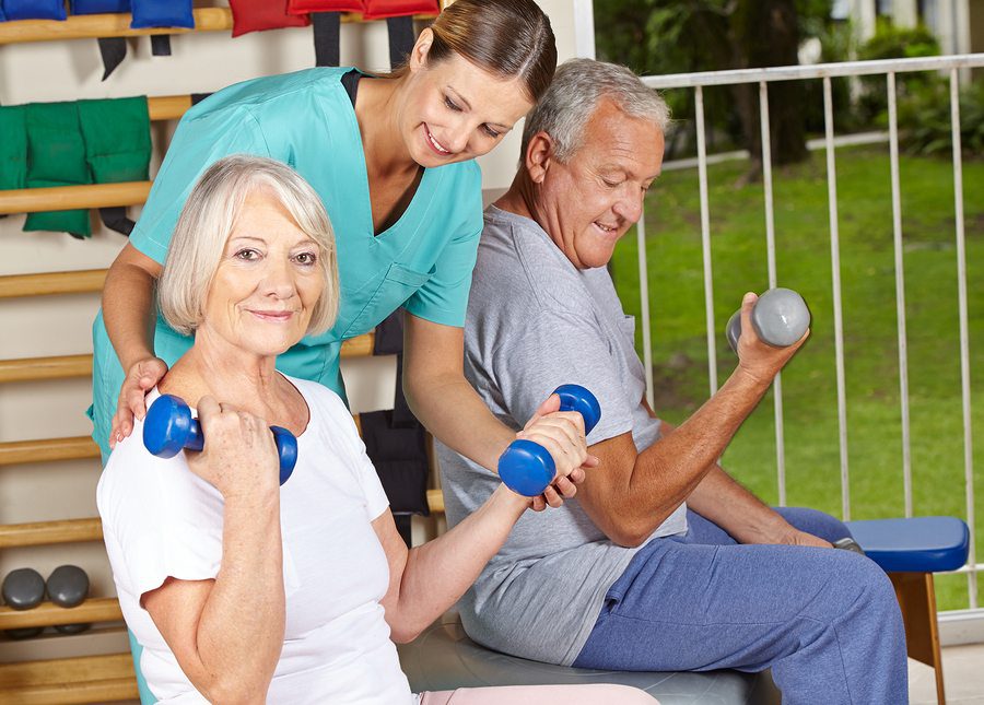Older woman and man lifting dumbbells during a physiotherapy session, assisted by a smiling therapist.