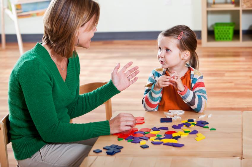 Speech therapist engaging with a young girl while working with colourful geometric shapes at a table.