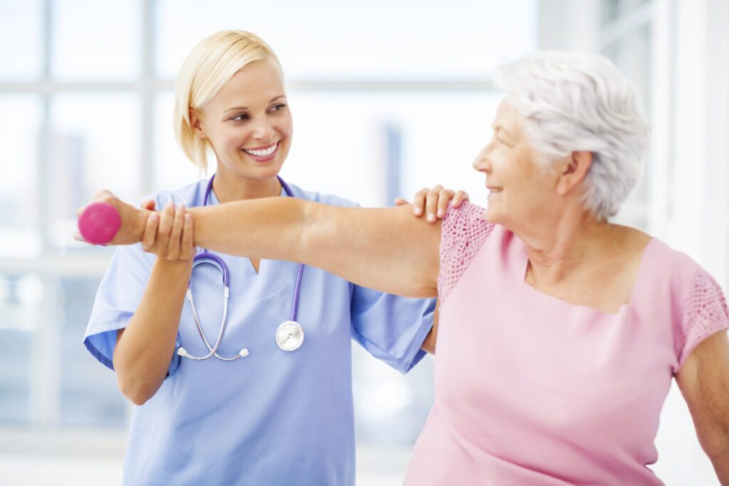 Smiling healthcare professional assisting an older woman with arm exercises using a small weight.