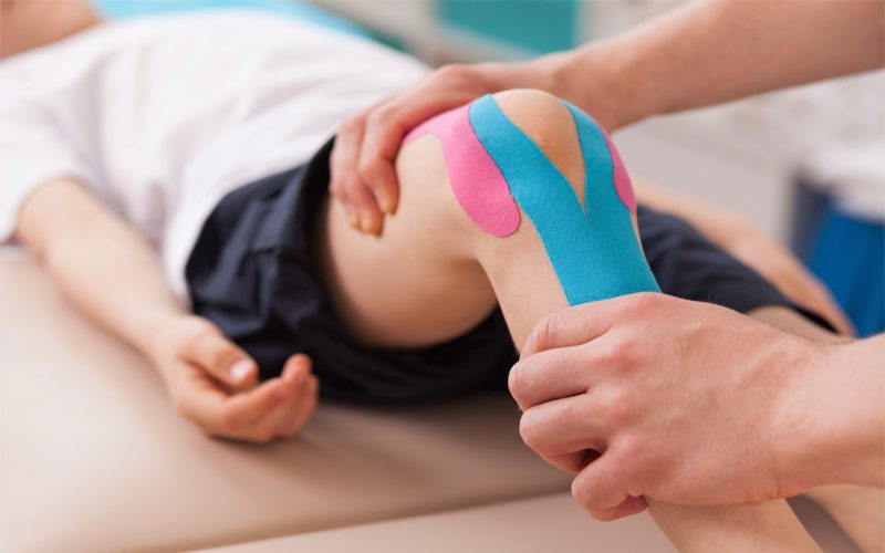 Physiotherapist applying colourful kinesiology tape to a patient’s knee during treatment.
