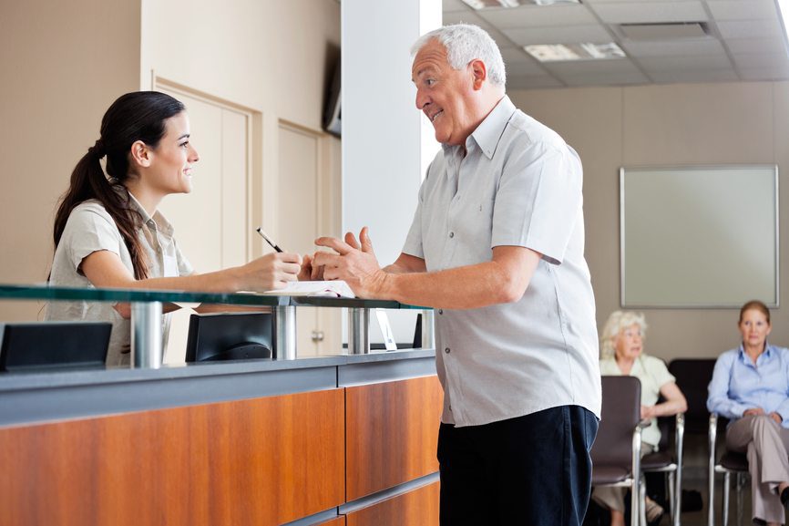 Friendly receptionist assisting senior patient at medical clinic front desk.